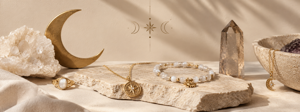 A styled flat-lay of gold-toned jewelry, including a necklace with a crescent moon pendant and a beaded bracelet with pearl-like beads and a small gold charm, arranged on a stone slab with crystals, a moon ornament, and a bowl in a warm neutral setting.